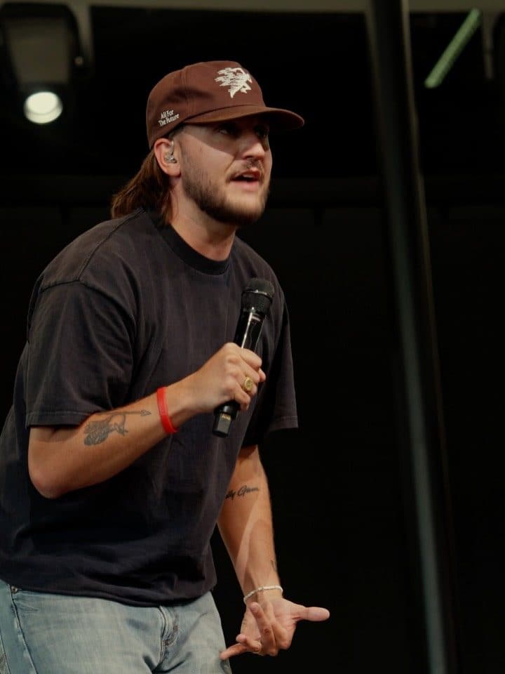 Chase on stage in a cap and black tee, a microphone in one hand, the other open